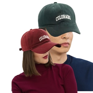 May include: Two baseball caps, one dark green and one burgundy, are displayed. The word "COLORADO" is embroidered in white on the front of each cap. The caps are worn by two people, one wearing a burgundy turtleneck and the other a navy blue shirt.