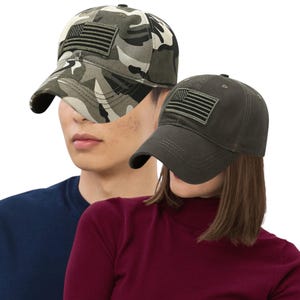 May include: Two baseball caps with an American flag patch. One cap has a camouflage pattern in shades of green, brown, and white. The other cap is solid olive green. Both caps have a curved brim and adjustable strap.