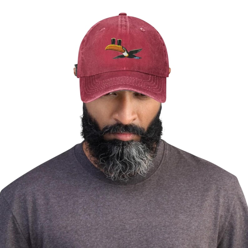 K&ouml;nnte beinhalten: Burgunderfarbene Baseballkappe mit einem gestickten Tukan-Design. Der Tukan hat einen gelben Schnabel, einen schwarz-wei&szlig;en K&ouml;rper und blaue Akzente. Die Kappe hat einen gebogenen Schirm und einen verstellbaren Riemen.