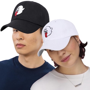 May include: Black and white baseball caps with a cartoon ghost design. The ghost has a white body, a red tongue, and a wide open mouth. The caps are made of a soft material and have a curved brim.