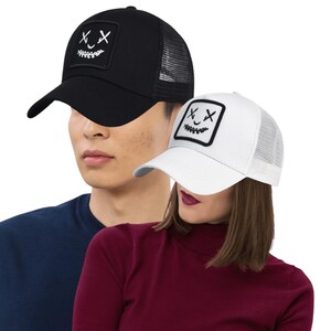 May include: Black and white trucker hats with a patch featuring a stitched face design. The black hat has a black front panel and brim with a mesh back. The white hat has a white front panel and brim with a mesh back.