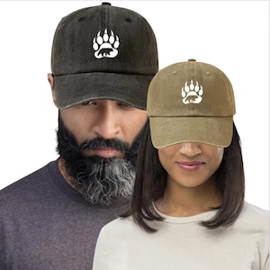 Peut inclure: Deux casquettes de baseball, une gris foncé et une kaki, sont présentées. Chaque casquette arbore une empreinte de patte d'ours blanche avec une silhouette d'ours. Les casquettes sont portées par deux personnes, l'une avec une barbe et l'autre avec des cheveux foncés.