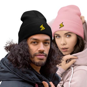 May include: Black and pink beanies featuring a yellow lightning bolt logo. The black beanie is worn by a person with long, curly hair and a black jacket. The pink beanie is worn by a person in a pink hoodie.