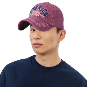 May include: A maroon baseball cap with the word "AMERICA" embroidered in navy blue arched across the front. A small patch with the American flag and the number "1776" is also on the front. The cap has a curved brim and white stitching.