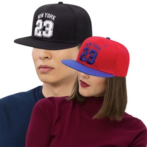 May include: Two baseball caps with flat brims. One is black with white text reading "NEW YORK 23". The other is red with a blue brim and blue text reading "NEW YORK 23". Both caps have a structured design.