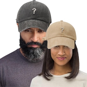 Peut inclure: Deux casquettes de baseball, une grise foncée et une beige, chacune avec un emblème de point d'interrogation argenté. Les casquettes ont un aspect vieilli. L'arrière-plan présente deux personnes.