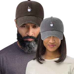 May include: Two baseball caps, one brown and one grey, each embroidered with a white coffee cup design. The caps are worn by two people. The caps are a casual accessory. The caps are a casual accessory.