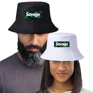 May include: Two bucket hats, one black and one white, are displayed. Each hat features the word "Savage" in white lettering, set against a black background with green cannabis leaf designs. The hats are shown on two people.