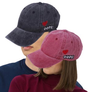 Peut inclure: Deux casquettes de baseball, une bleu foncé et une bordeaux, sont présentées. Chaque casquette arbore un écusson cœur rouge et le mot "OOPS!" brodé sur le côté. Les casquettes ont un aspect classique et légèrement usé.
