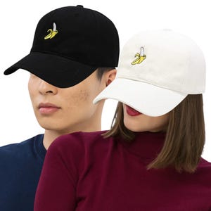 May include: Black and white baseball caps, each with a yellow banana embroidered on the front. The caps have a curved peak and a classic six-panel design. The black cap is worn by a person wearing a blue top, and the white cap is worn by a person wearing a burgundy top.