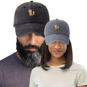 May include: Two gray baseball caps with a fox embroidery. The caps are made of a soft, washed fabric. The fox is orange and white. The caps are a casual accessory.