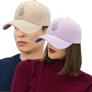 May include: Two baseball caps, one tan and one lavender, each featuring an embroidered peace sign. The caps have a curved brim and adjustable strap. The tan cap is worn by a person wearing a navy blue shirt, and the lavender cap is worn by a person wearing a burgundy turtleneck.