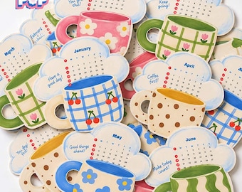 May include: A collection of cloud-shaped paper calendars featuring colorful mug illustrations with patterns like cherries, flowers, and polka dots. Each calendar displays a month, dates, and motivational phrases like You got this and Coffee first.