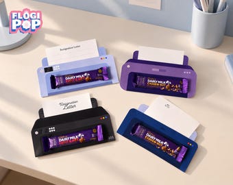 May include: Four printer-shaped paper holders in light purple, dark purple, black, and blue. Each holder features a slot for a white card and a horizontal opening displaying a Cadbury Dairy Milk chocolate bar. The logo FLOGI POP is visible.