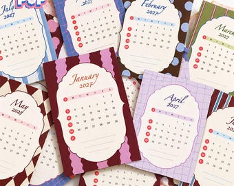 May include: A collection of 2027 calendar cards featuring the brand name FLOGI POP. Each card displays a specific month with a unique patterned background, including stripes, polka dots, and grids, surrounding a central scalloped white frame.