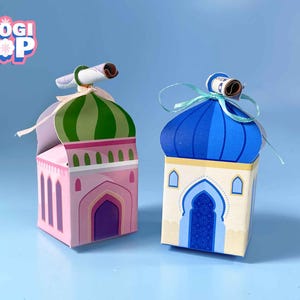 Puede incluir: Dos cajas decorativas con forma de mezquita. Una es rosa con una cúpula a rayas verdes y blancas, la otra es crema con una cúpula azul. Ambas tienen un pergamino en la parte superior y están atadas con una cinta. Las cajas están etiquetadas "FLOGI POP".