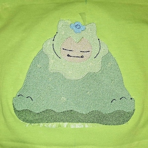 Könnte beinhalten: Ein lindgrünes T-Shirt mit einer zentralen Stickerei, die ein stilisiertes Wesen mit einer grünen Kapuze und einer kleinen blauen Blumenapplikation zeigt. Das Wesen hat geschlossene Augen und einen einfachen Mund auf grünem Grund.