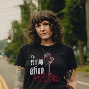 May include: Black t-shirt with the text "I'm Coming Alive" and a graphic of a red hand rising from the ground. The shirt is short-sleeved and has a crew neck.