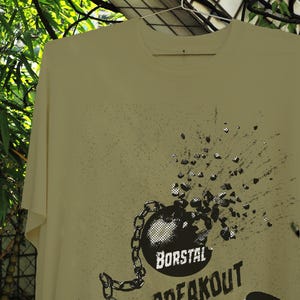 May include: A light yellow t-shirt with a graphic design featuring a black ball and chain with the words "BORSTAL BREAKOUT". The design includes a chain and a broken ball. The shirt is hanging on a hanger.