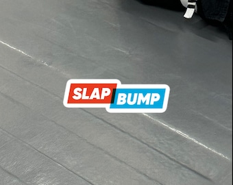 Puede incluir: Una estera gris con las palabras "SLAP BUMP" en una pegatina blanca, roja y azul. La pegatina es rectangular con esquinas redondeadas. La estera tiene líneas visibles y parece estar en un gimnasio o área de entrenamiento.