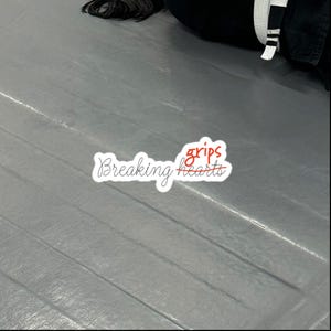 Pode incluir: Um autocolante branco com as palavras "Breaking grips hearts" em itálico. A palavra "grips" é vermelha, e a palavra "hearts" tem um traço a atravessá-la. O autocolante está numa superfície cinzenta.