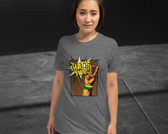 Puede incluir: Camiseta gris jaspeado con la frase "THAT'S TWO!" en un bocadillo de cómic. El gráfico incluye una mano con una pulsera verde y amarilla haciendo el signo de la victoria. La camiseta es de manga corta.