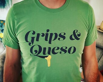 Puede incluir: Camiseta verde con el texto "Grips & Queso" en escritura azul marino. Un gráfico de queso amarillo está debajo del texto. La camiseta tiene mangas cortas y cuello redondo.