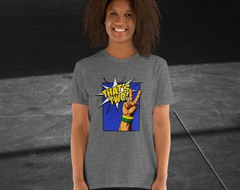 Puede incluir: Una camiseta gris con un gráfico de estilo cómic que dice "THAT'S TWO!" con una mano haciendo el signo de la paz. La mano tiene una pulsera verde y amarilla. La camiseta la lleva una persona.