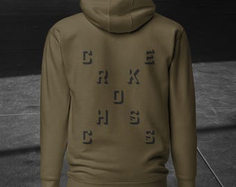 Puede incluir: Sudadera con capucha verde oliva con la palabra "SKETCHES" impresa en gris oscuro en la espalda. La sudadera tiene capucha y mangas largas. El fondo es gris oscuro y negro.