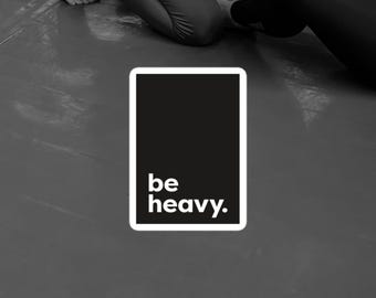 May include: A black and white graphic with the words "be heavy." The text is in a white font, set against a black rectangle with a white border. The background is a gray surface, with parts of people in the background.