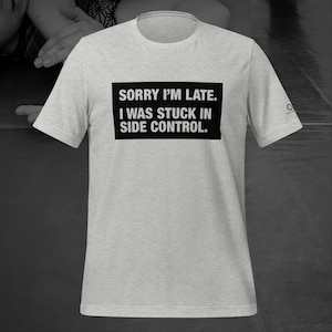 May include: A light gray t-shirt with the text "SORRY I'M LATE. I WAS STUCK IN SIDE CONTROL." in a black rectangle. The shirt has short sleeves and a crew neck. A small logo is visible on the sleeve.
