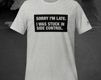 Puede incluir: Una camiseta gris claro con el texto "SORRY I'M LATE. I WAS STUCK IN SIDE CONTROL." en un rectángulo negro. La camiseta tiene mangas cortas y cuello redondo. Un pequeño logotipo es visible en la manga.