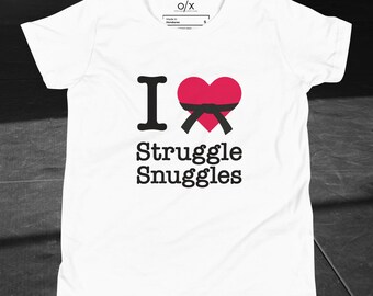 Puede incluir: Camiseta blanca con el texto "I ❤️ Struggle Snuggles" en fuente negra. El corazón es rojo con un diseño de cinturón negro. La camiseta tiene cuello redondo y mangas cortas.
