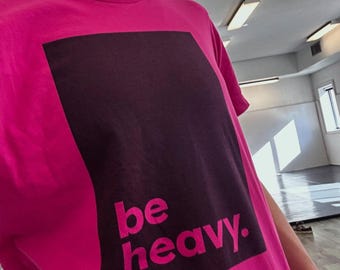 Puede incluir: Camiseta magenta con un diseño rectangular morado oscuro en la parte delantera. Las palabras "be heavy." están impresas en magenta debajo. La camiseta tiene mangas cortas y cuello redondo.