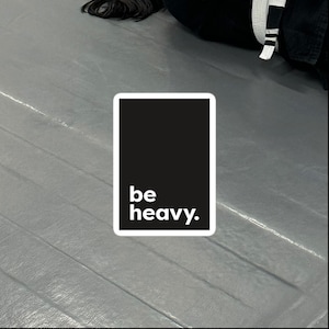 May include: A rectangular sticker with a black background and white border. The words "be heavy." are printed in white, sans-serif font. The sticker is on a gray surface.