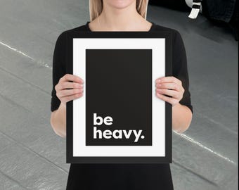 Puede incluir: Una impresión en blanco y negro enmarcada con las palabras "be heavy." en negrita, fuente sans-serif. La impresión tiene un borde blanco y es sostenida por una persona que lleva una parte superior negra.