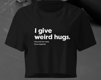 Puede incluir: Top corto negro con el texto blanco "I give weird hugs." y las frases "Around the neck. From behind." debajo. La camiseta tiene mangas cortas y cuello redondo.