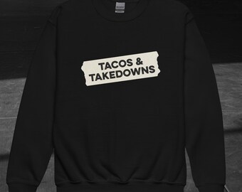 Puede incluir: Sudadera negra con la frase "TACOS & TAKEDOWNS" en blanco desgastado. El texto está impreso en una forma rectangular blanca. El logotipo de Gildan es visible en el cuello. Prenda casual y cómoda.