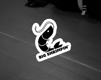 May include: A black and white sticker featuring a stylized graphic of a shrimp with a curved tail and the text "BIG SHRIMPIN'" printed in bold, sans-serif capital letters. The design has a thick white border around the black illustration.