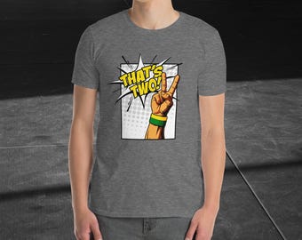 Puede incluir: Camiseta gris con un gráfico estilo cómic. El gráfico muestra una mano haciendo el signo de la paz con el texto "THAT'S TWO!" en un bocadillo amarillo y blanco. La mano tiene una pulsera amarilla y una pulsera verde.