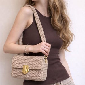 Könnte beinhalten: Eine beige gehäkelte Handtasche mit rechteckiger Struktur und strukturiertem Strickmuster. Die Tasche verfügt über einen dicken, passenden Häkel-Schulterriemen und einen polierten goldfarbenen Metalldruckverschluss an der Vorderklappe.