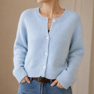 Könnte beinhalten: Eine hellblaue Strickjacke mit Rundhalsausschnitt, langen Ärmeln mit gerippten Bündchen und einer Knopfleiste mit vier weißen Knöpfen. Das Kleidungsstück hat einen gerippten Saum und wird über einer blauen Jeans mit einem braunen Ledergürtel getragen.