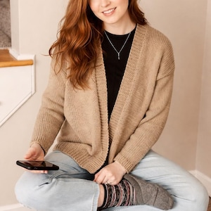 Peut inclure: Une femme porte un cardigan en tricot beige, un chemisier noir et un jean bleu clair. Elle est assise sur une surface recouverte de moquette, tenant un smartphone noir. Ses chaussettes ont un motif. L'arrière-plan est de couleur neutre.