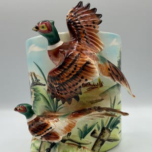 May include: A cylindrical ceramic vase featuring a hand-painted landscape with two pheasants. One pheasant is depicted in flight with brown, black, and green plumage, while the other stands on the ground below among green grass and foliage.