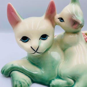 May include: A ceramic figurine of a mother cat and kitten. The cats are cream-colored with green and pink accents. The mother cat is lying down, and the kitten is nestled on top. The eyes are blue, and the nose is black.