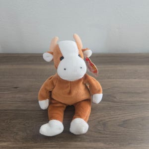 Puede incluir: Un peluche de vaca con pelaje marrón y blanco, con pequeños cuernos y una etiqueta. La vaca tiene patas blancas y una mancha blanca en la cara. El juguete está sobre una superficie de madera.
