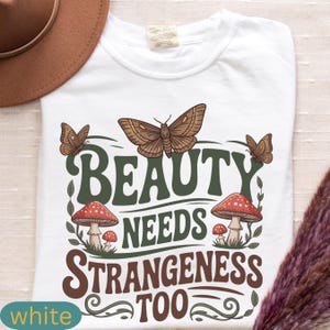 Może przedstawiać: Biała koszulka z graficznym wzorem przedstawiającym motyle, grzyby i napis "BEAUTY NEEDS STRANGENESS TOO" w kolorze zielonym i brązowym. Koszulka ma okrągły dekolt i kr&oacute;tkie rękawy. Brązowy kapelusz widoczny w lewym g&oacute;rnym rogu.