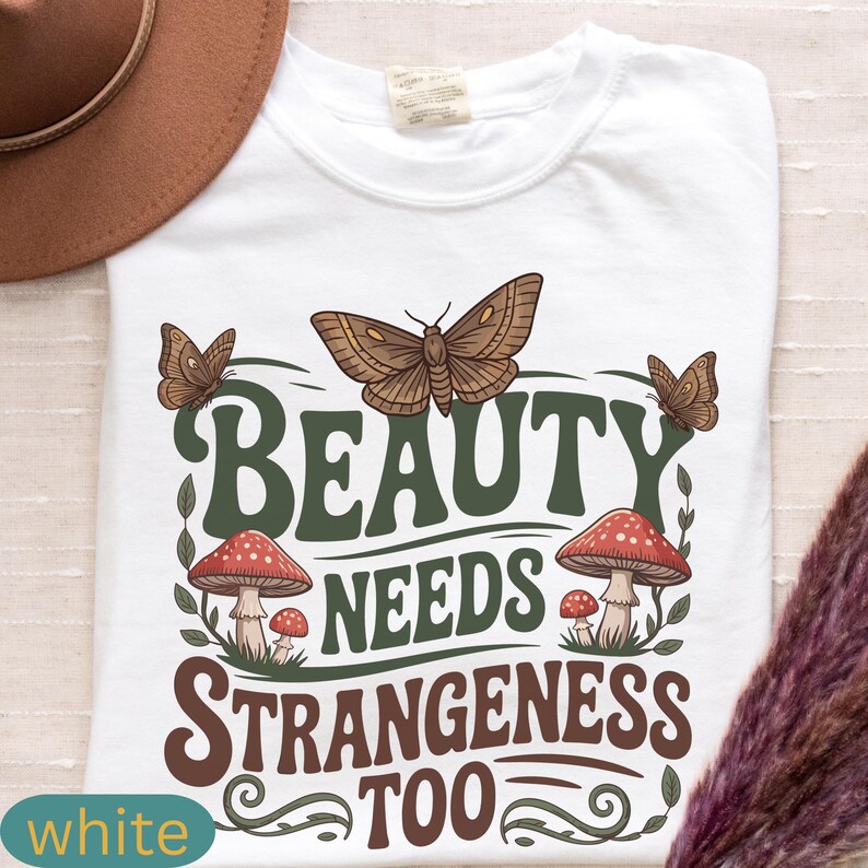 Może przedstawiać: Biała koszulka z graficznym wzorem przedstawiającym motyle, grzyby i napis "BEAUTY NEEDS STRANGENESS TOO" w kolorze zielonym i brązowym. Koszulka ma okrągły dekolt i kr&oacute;tkie rękawy. Brązowy kapelusz widoczny w lewym g&oacute;rnym rogu.