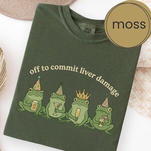 May include: A moss green t-shirt featuring a graphic of four frogs wearing party hats and a crown, holding various alcoholic drinks. The text "off to commit liver damage" is printed in a curved arch above the frogs in a light beige font.