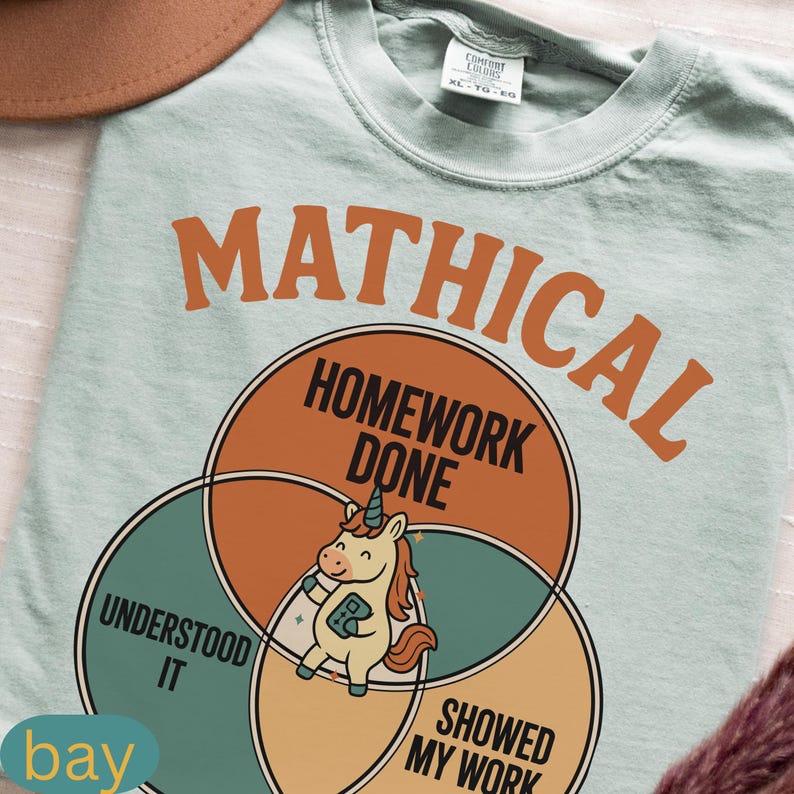 Peut inclure: Un t-shirt bleu clair avec le mot "MATHICAL" en lettres orange. En dessous, un diagramme de Venn montre "HOMEWORK DONE" avec une licorne de dessin anim&eacute;. Les cercles indiquent "UNDERSTOOD IT" et "SHOWED MY WORK". Le mot "bay" est en bas.